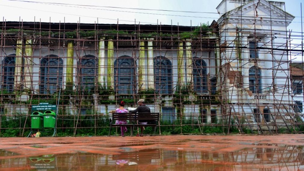 Nepali people sit on a bench in front of a royal palace covered by scaffolding as it undergoes repairs in Durbar Square in Kathmandu 