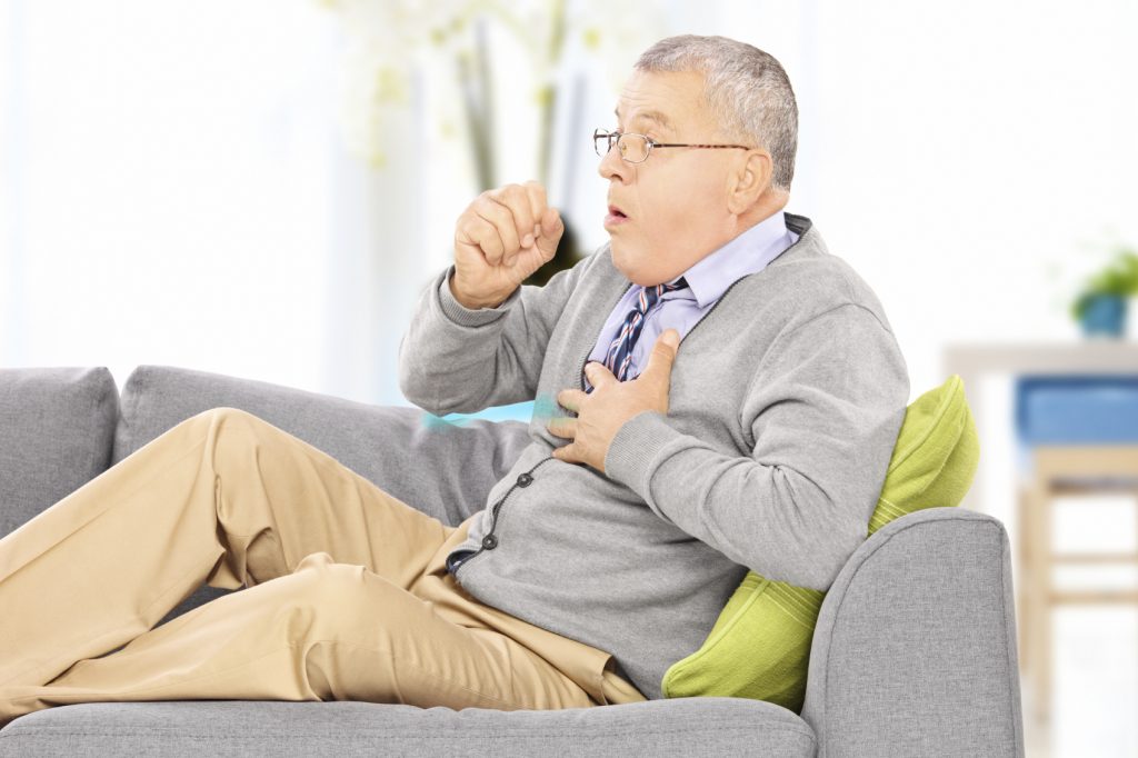 Mature man seated on a sofa coughing because of pulmonary disease at home